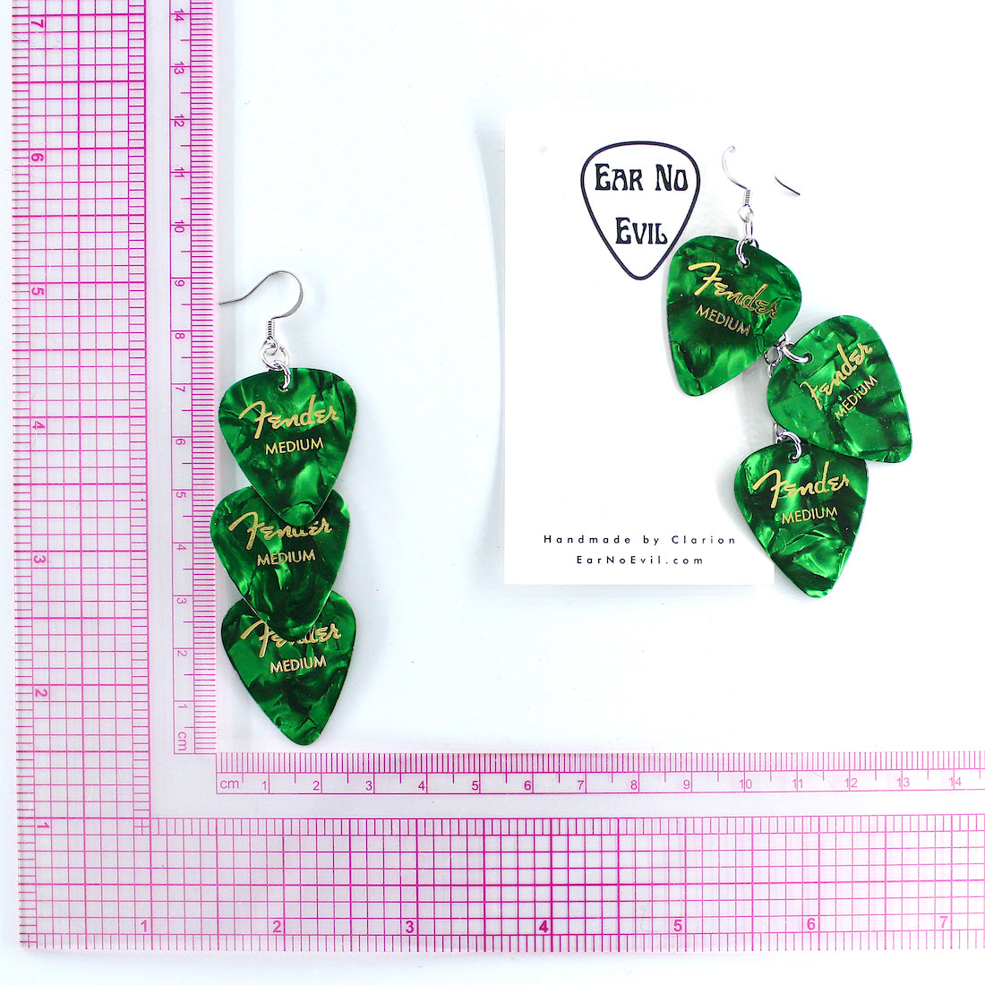 A pair of Genuine green Fender guitar-pick waterfall shaped dangle earrings with silver, hypoallergenic stainless steel fish hook earring wires. The earrings are displayed on a white background with a, Ear No Evil brand card, and a pink grid ruler to show the drop height of the earring is 3.5 inches.