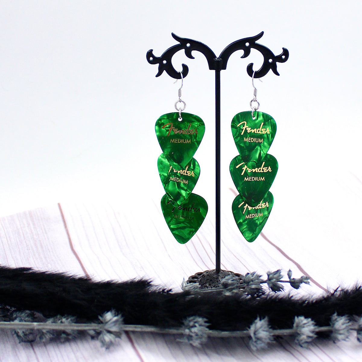 Three Authentic Emerald Green Fender guitar picks in a tiered drop shaped earring with silver, hypoallergenic, stainless steel fish hook earring wires.  The earrings are displayed on a Punk rock inspired black stand against a light gray background. 