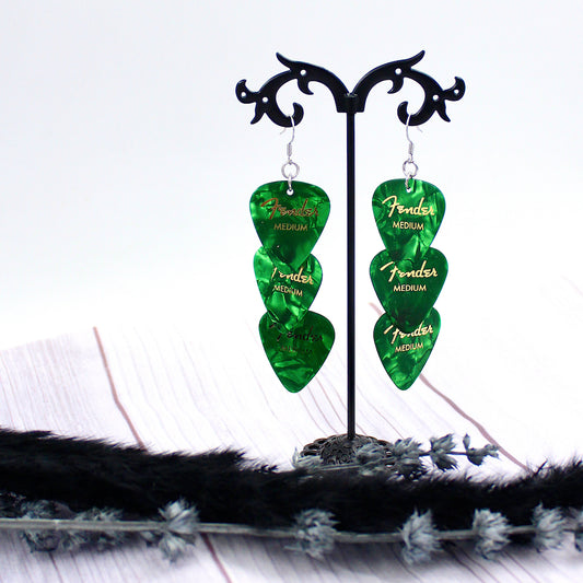 Three Authentic Emerald Green Fender guitar picks in a tiered drop shaped earring with silver, hypoallergenic, stainless steel fish hook earring wires.  The earrings are displayed on a Punk rock inspired black stand against a light gray background. 