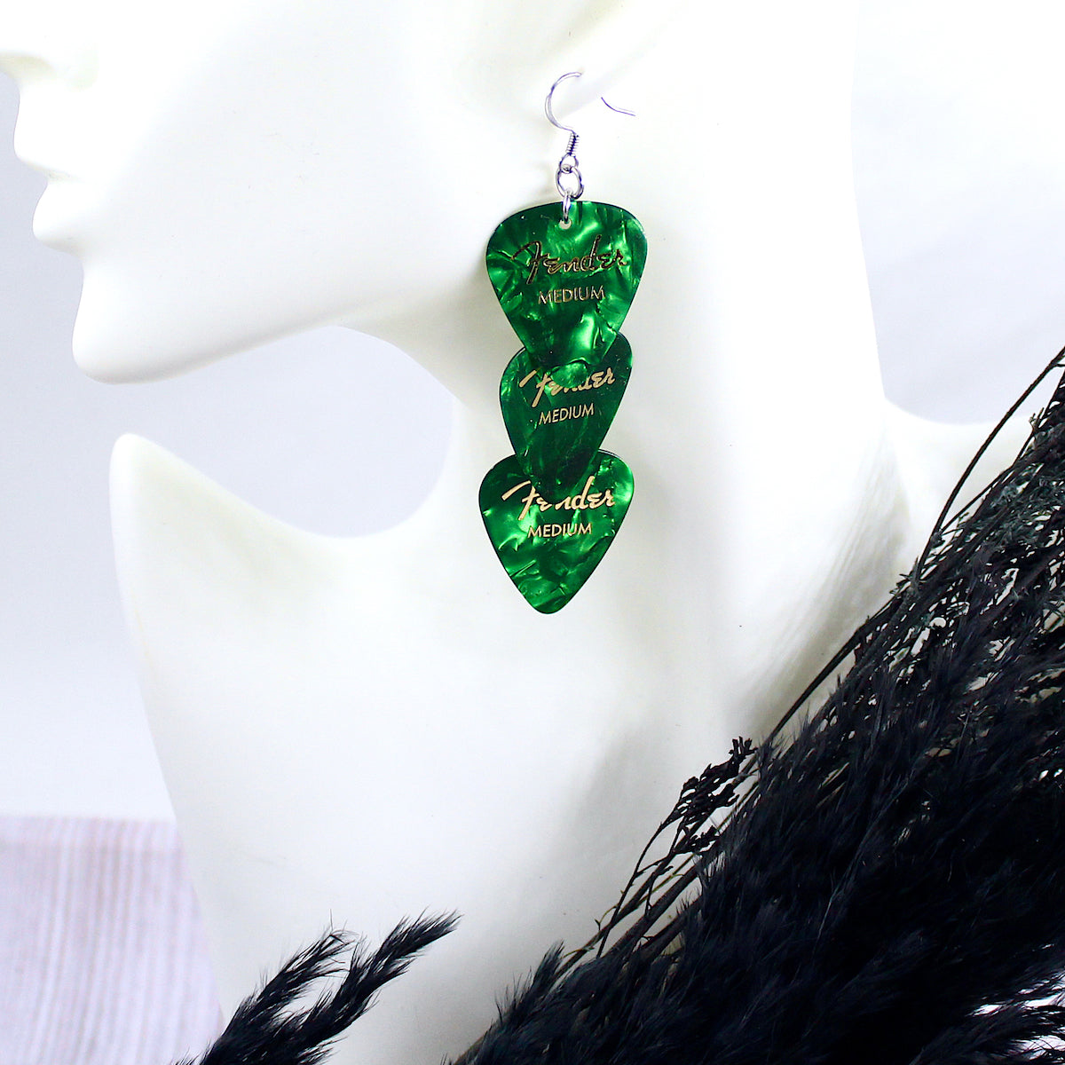 Three Green authentic Fender guitar picks strung vertically in an earring on a white mannequin head, with hypo allergenic, silver, stainless steel fishhook ear wires. The deep green picks are printed with gold Fender logos and 'MEDIUM'. 