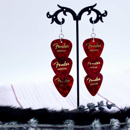 Three Authentic Red Fender guitar picks in a tiered drop shaped earring with silver, hypoallergenic, stainless steel fish hook earring wires.  The earrings are displayed on a Punk rock inspired black stand against a light gray background.