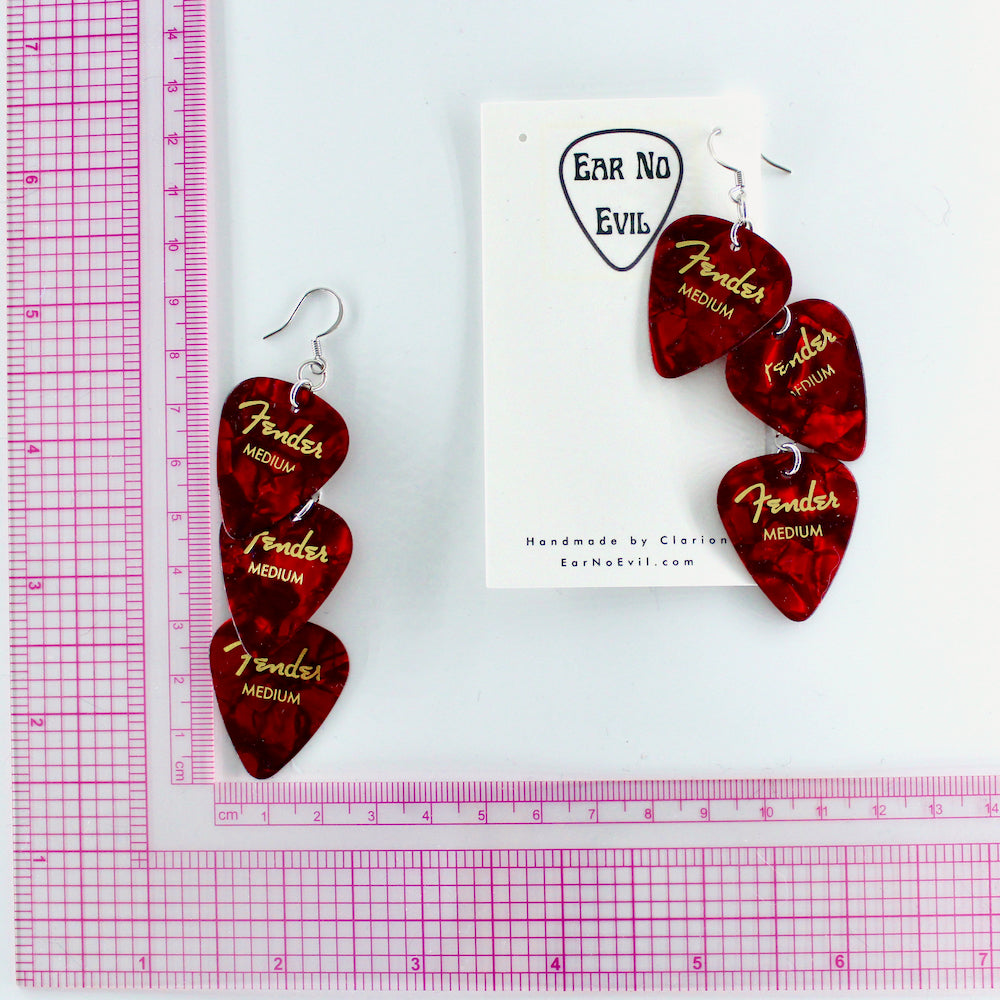 A pair of red Fender guitar-pick waterfall shaped dangle earrings with silver, hypoallergenic stainless steel fish hook earring wires.  The earrings are displayed on a white background with a, Ear No Evil brand card, and a pink grid ruler to show the drop height of the earring is 3.5 inches.
