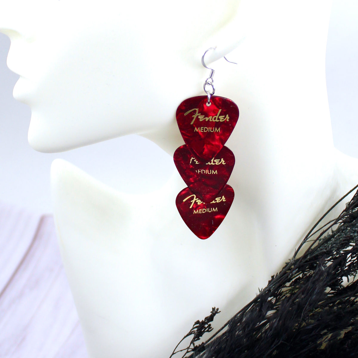 Three red authentic Fender guitar picks strung vertically in an earring on a white mannequin head, with hypo allergenic, silver, stainless steel fishhook ear wires. The deep red picks are printed with gold Fender logos and 'MEDIUM'.