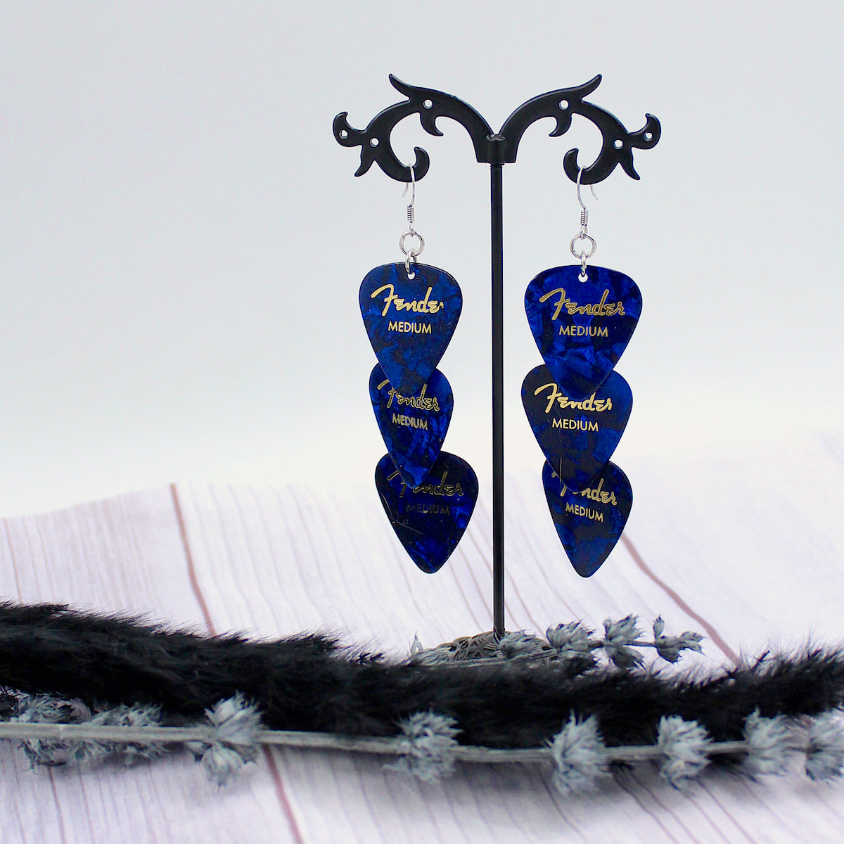 Three Authentic sapphire blue Fender guitar picks in a tiered drop shaped earring with silver, hypoallergenic, stainless steel fish hook earring wires.  The earrings are displayed on a Punk rock inspired black stand against a light gray background. 