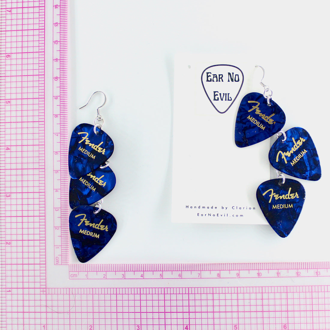 A pair of genuine blue Fender guitar-pick waterfall shaped dangle earrings with silver, hypoallergenic stainless steel fish hook earring wires.  The earrings are displayed on a white background with a, Ear No Evil brand card, and a pink grid ruler to show the drop height of the earring is 3.5 inches. 