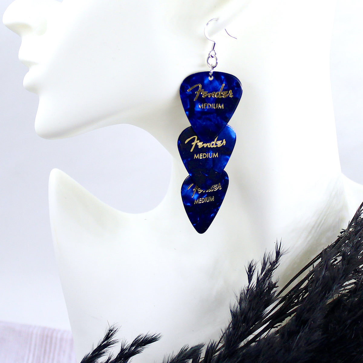 Three deep blue authentic Fender guitar picks strung vertically in an earring on a white mannequin head, with hypo allergenic, silver, stainless steel fishhook ear wires. The deep blue picks are printed with gold Fender logos and 'MEDIUM'. 